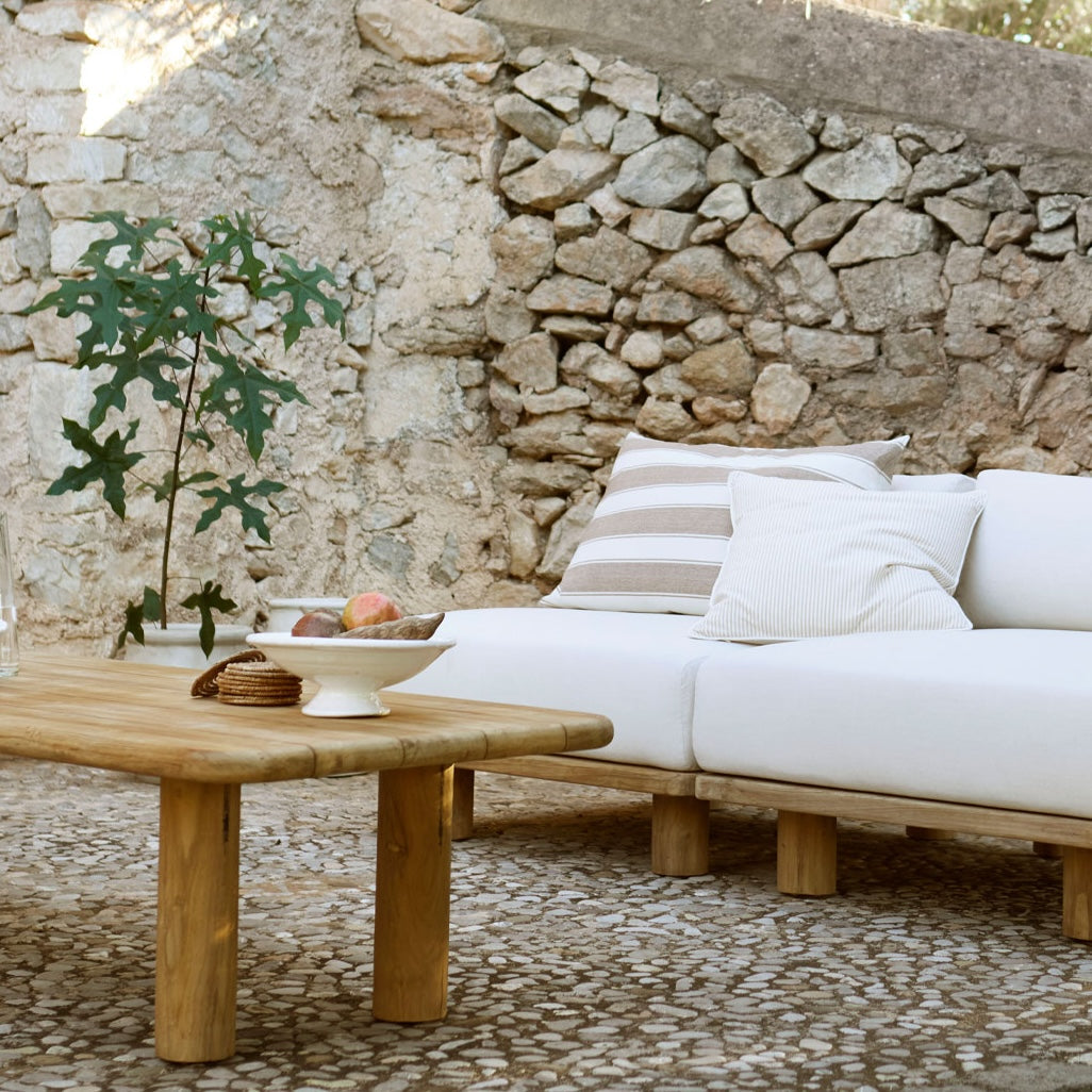 Outdoor setting with a wooden table and white cushions against a stone wall.