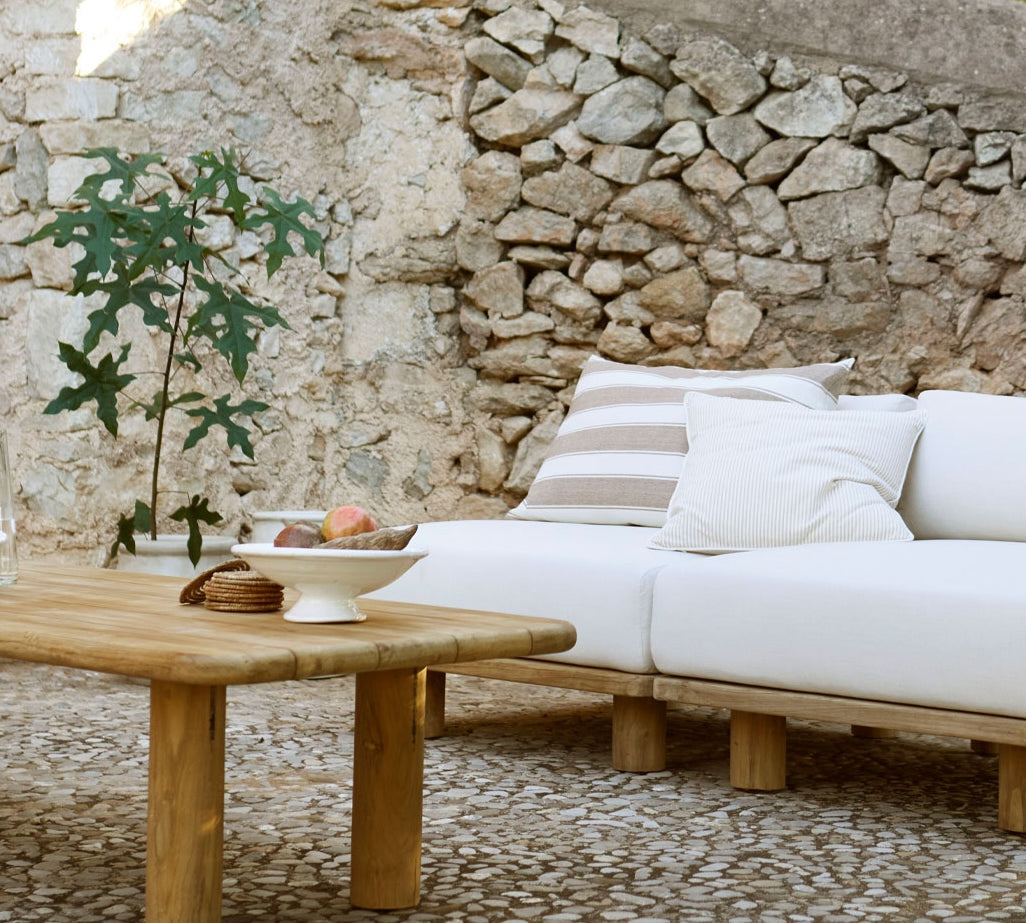 Outdoor setting with a wooden table and white cushions against a stone wall.