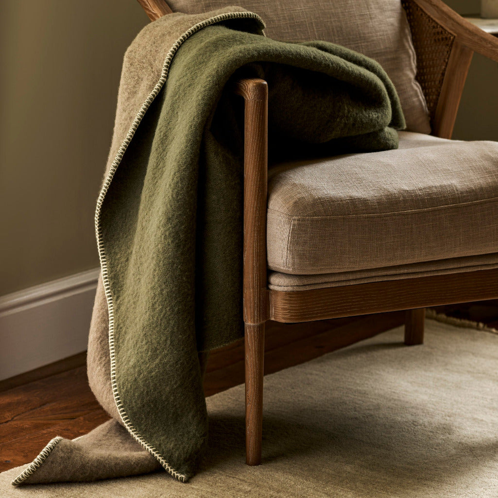A cozy olive green throw blanket with a cream fringed edge draped over a mid-century modern armchair with beige upholstery, set in a warmly lit living room.