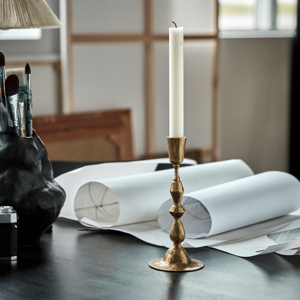 Gold candlestick with a white candle on a table with art supplies and papers.