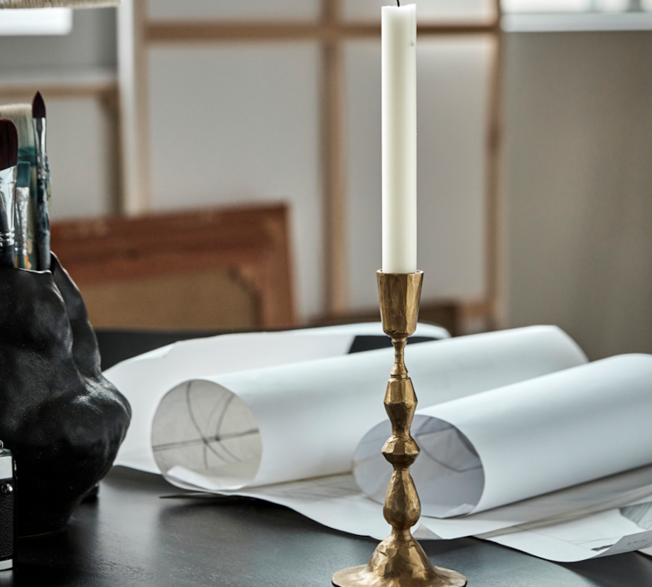 Gold candlestick with a white candle on a table with art supplies and papers.