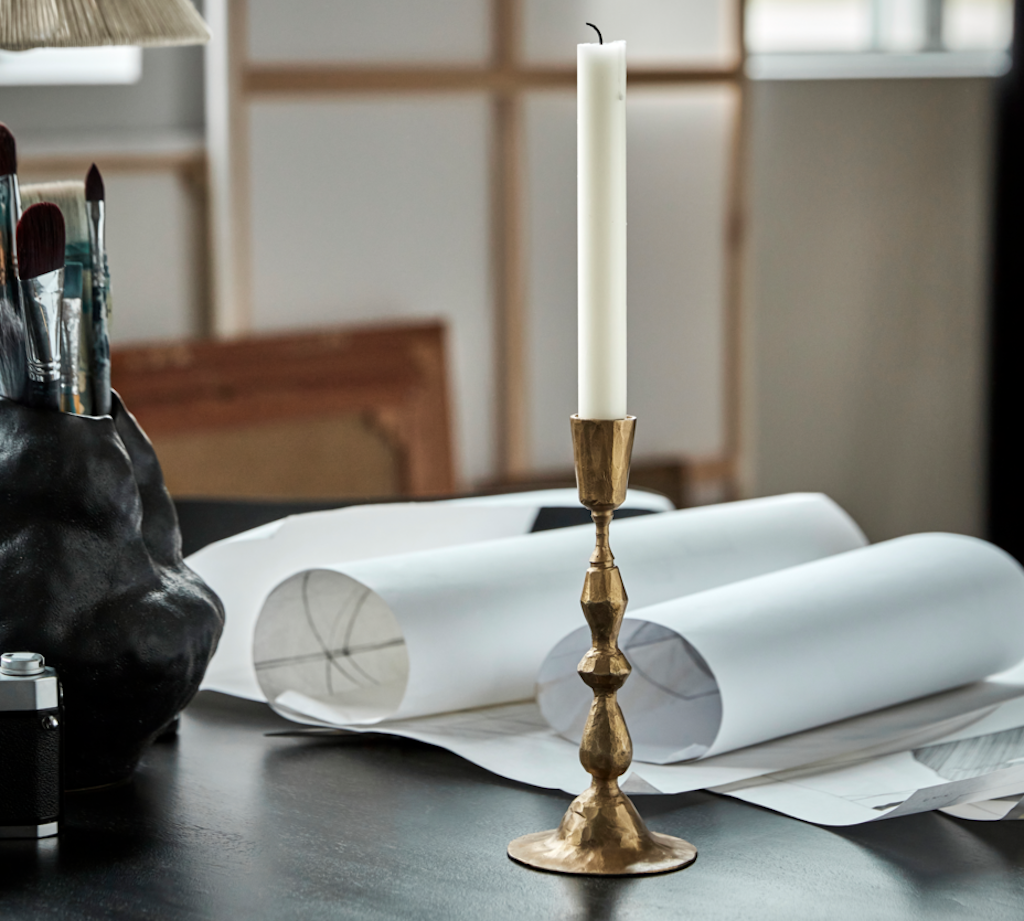Gold candlestick with a white candle on a table with art supplies and papers.