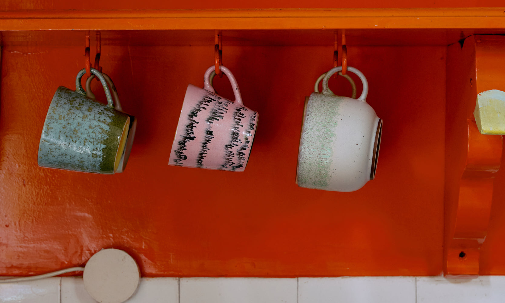 Three mugs hanging on an orange shelf with a tiled wall and blue shelf in the background.