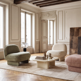 Three modern armchairs in sage green, cream, and dusty pink arranged around a low cream coffee table on a light rug in a classic Parisian-style room with wood paneling and herringbone parquet flooring.