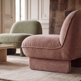 A close-up of a plush, dusty rose velvet armchair in a beautifully styled living room with a beige coffee table, a light rug, and a second muted green armchair in the background.