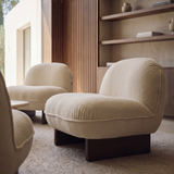 A modern curved upholstered armchair in a warm cream bouclé fabric with dark wooden legs, shown in a stylishly decorated living room with other matching chairs and a wooden slatted wall.
