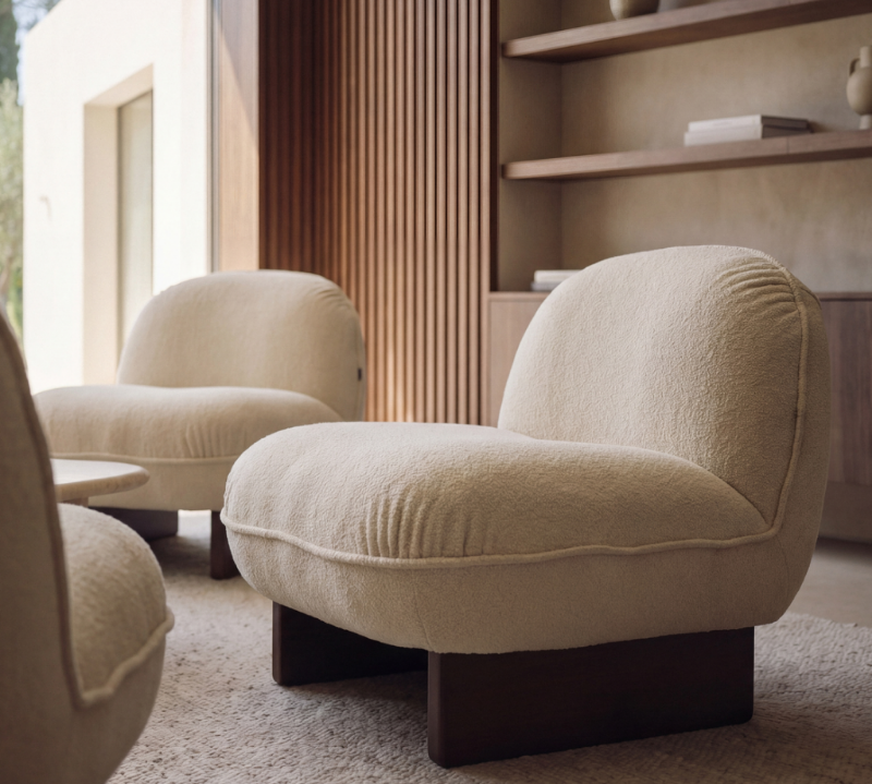 A modern curved upholstered armchair in a warm cream bouclé fabric with dark wooden legs, shown in a stylishly decorated living room with other matching chairs and a wooden slatted wall.