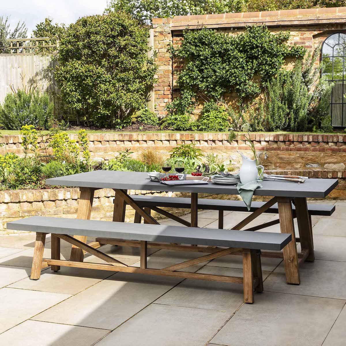 A Chilford dining table with a gray concrete top and acacia wood frame and legs, placed outdoors on a patio with garden in the background.