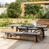A Chilford dining table with a gray concrete top and acacia wood frame and legs, placed outdoors on a patio with garden in the background.