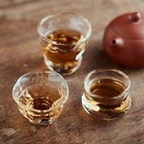 Three small glass cups filled with a amber liquid on a wooden surface, with a red teapot in the background.