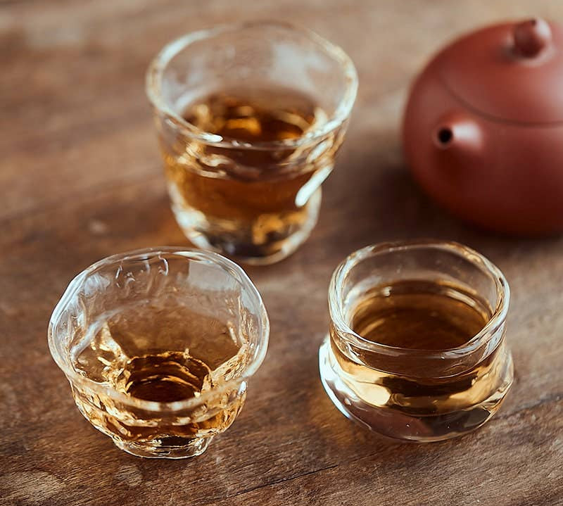 Three small glass cups filled with a amber liquid on a wooden surface, with a red teapot in the background.