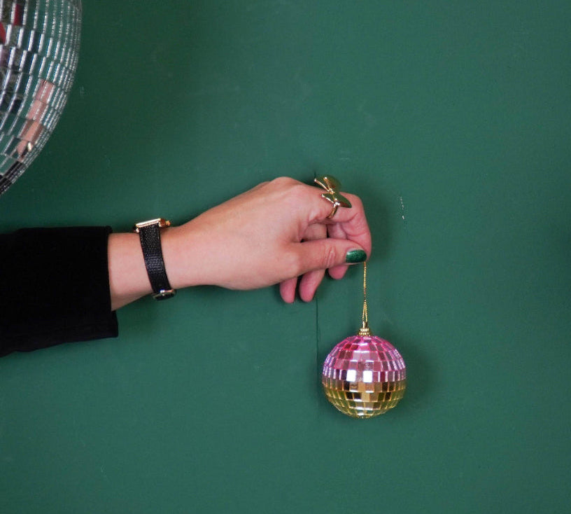 Hand holding a disco ball ornament against a green background with Christmas tree branches.