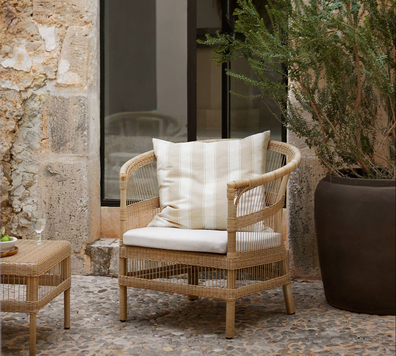 rattan chair with white cushion in an outdoor setting with stone wall and plant.