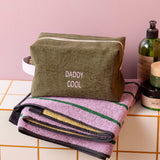 An olive green zippered cosmetic bag with 'DADDY COOL' embroidered on the front, resting on top of a folded lavender towel with green and gold stripes. The scene is styled on a tiled counter with bath bottles against a pink wall.