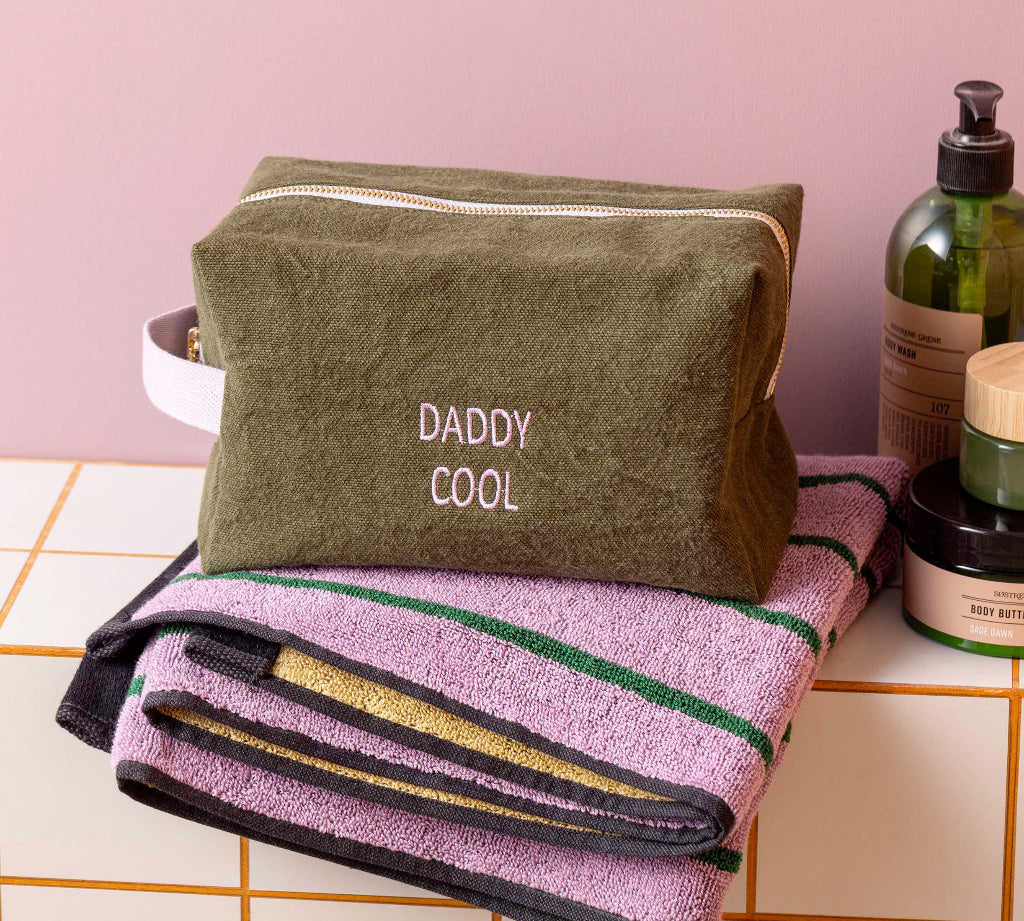 An olive green zippered cosmetic bag with 'DADDY COOL' embroidered on the front, resting on top of a folded lavender towel with green and gold stripes. The scene is styled on a tiled counter with bath bottles against a pink wall.