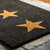 A dark charcoal gray rectangular doormat with two large tan stars, shown on a stone floor with a light rug border and shoes in the background.