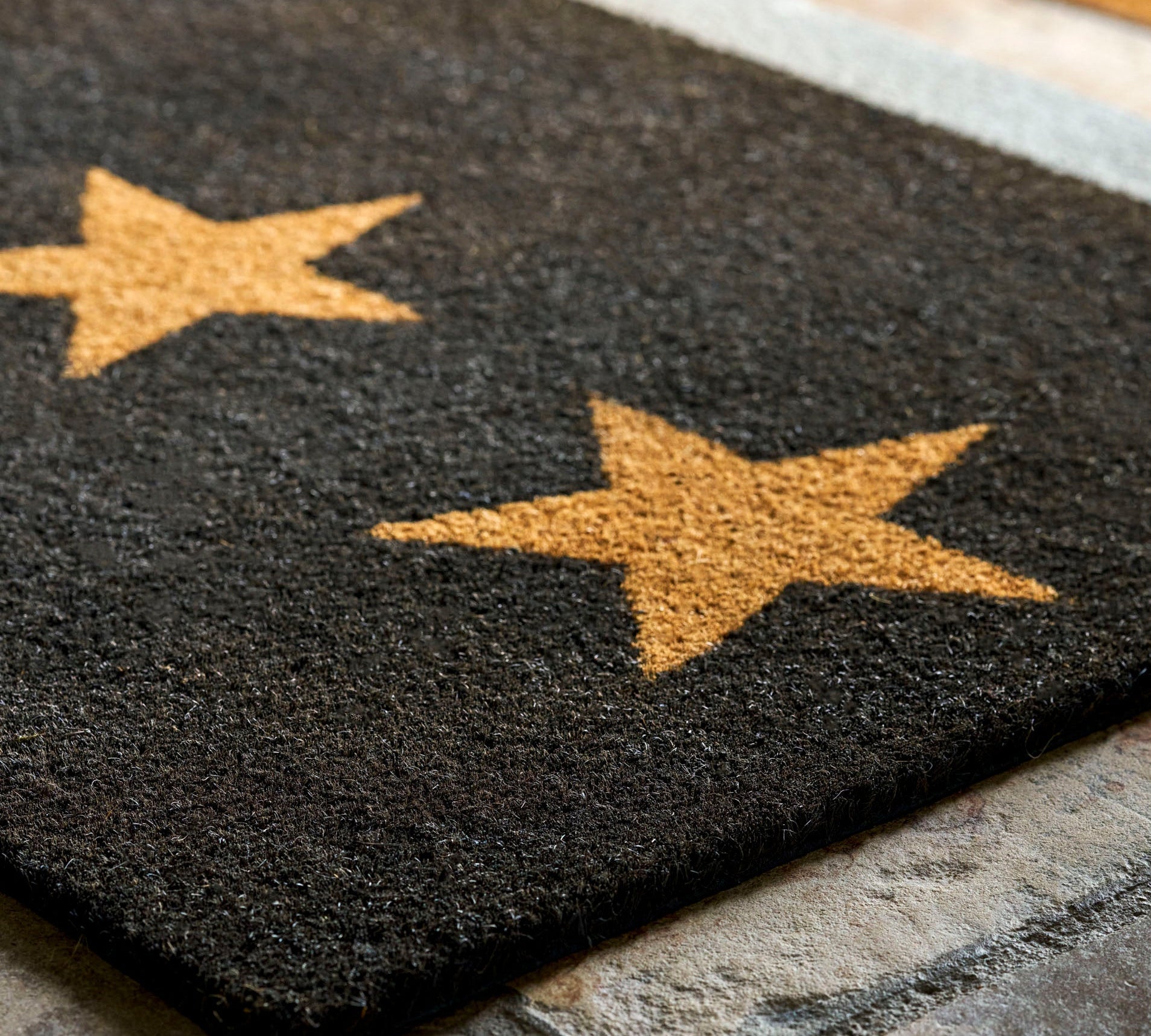A dark charcoal gray rectangular doormat with two large tan stars, shown on a stone floor with a light rug border and shoes in the background.