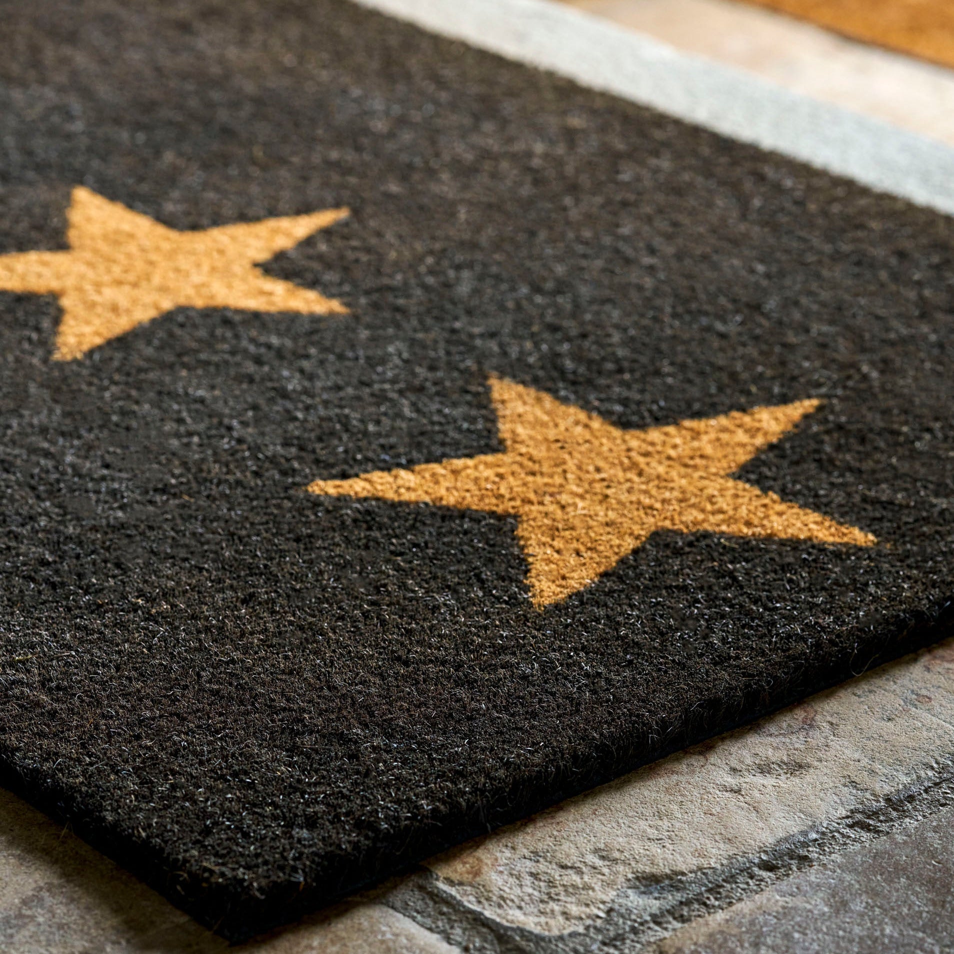 A dark charcoal gray rectangular doormat with two large tan stars, shown on a stone floor with a light rug border and shoes in the background.