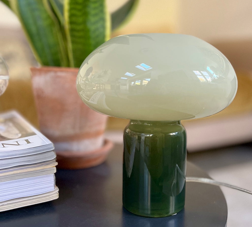 Green mushroom-shaped lamp on a table with books and a plant in the background