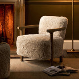 A cozy living room scene featuring a plush, cream-colored shaggy armchair with wooden arms, positioned in front of a fireplace. A matching shaggy ottoman is partially visible in the foreground, and a brass floor lamp with a beige shade stands to the right.