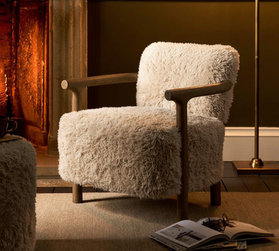 A cozy living room scene featuring a plush, cream-colored shaggy armchair with wooden arms, positioned in front of a fireplace. A matching shaggy ottoman is partially visible in the foreground, and a brass floor lamp with a beige shade stands to the right.