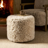 A round, shaggy off-white pouf ottoman styled in a cozy living room setting next to a matching armchair and in front of a lit fireplace.