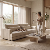 A beige upholstered sofa in a bright, modern living room with a dog resting on it, a woman standing nearby holding a mug, and a wooden coffee table in front.
