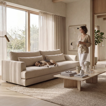 A beige upholstered sofa in a bright, modern living room with a dog resting on it, a woman standing nearby holding a mug, and a wooden coffee table in front.