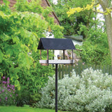 Bird feeder on a stand in a garden setting with greenery and flowers.