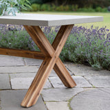 Outdoor dining table with a rectangular light gray concrete-look top and a rustic brown wooden X-shaped base, set on a stone patio with lavender and greenery in the background.
