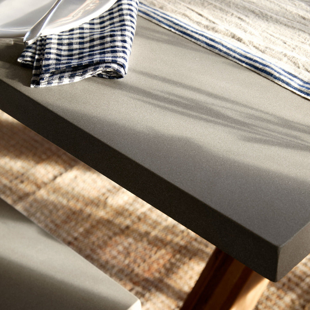 A close-up of a modern gray concrete dining table with a dark gray side edge, styled with a white plate, a blue-and-white gingham napkin, a blue-and-white striped runner, and a woven natural-fiber rug underneath.