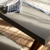A close-up of a modern gray concrete dining table with a dark gray side edge, styled with a white plate, a blue-and-white gingham napkin, a blue-and-white striped runner, and a woven natural-fiber rug underneath.