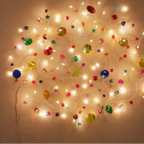 Warm white Christmas string lights with assorted colorful ornaments, including round baubles, stars, and snowflake shapes, arranged in a circular pattern against a plain beige wall.