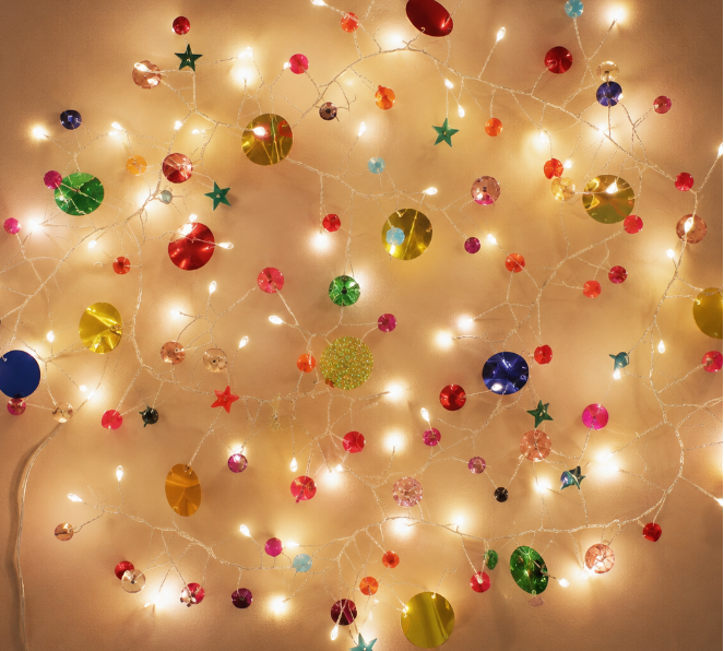 Warm white Christmas string lights with assorted colorful ornaments, including round baubles, stars, and snowflake shapes, arranged in a circular pattern against a plain beige wall.