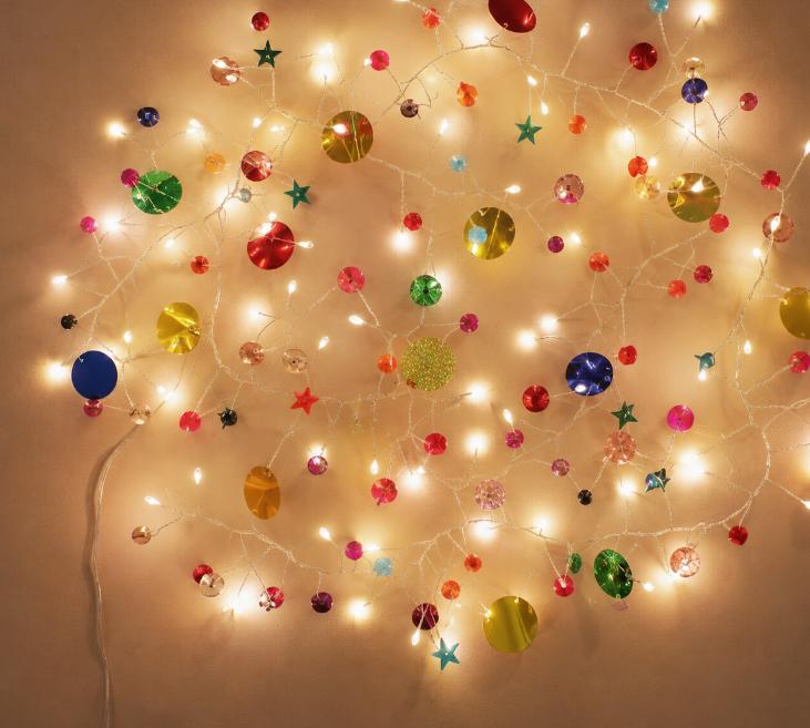 Warm white Christmas string lights with assorted colorful ornaments, including round baubles, stars, and snowflake shapes, arranged in a circular pattern against a plain beige wall.
