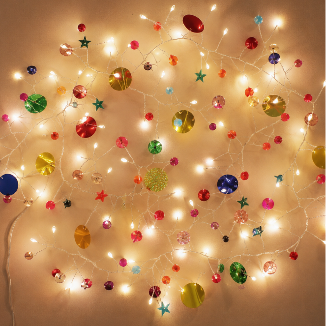 Warm white Christmas string lights with assorted colorful ornaments, including round baubles, stars, and snowflake shapes, arranged in a circular pattern against a plain beige wall.