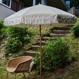 Beige striped patio umbrella with tassels on a wooden stand in a garden setting.