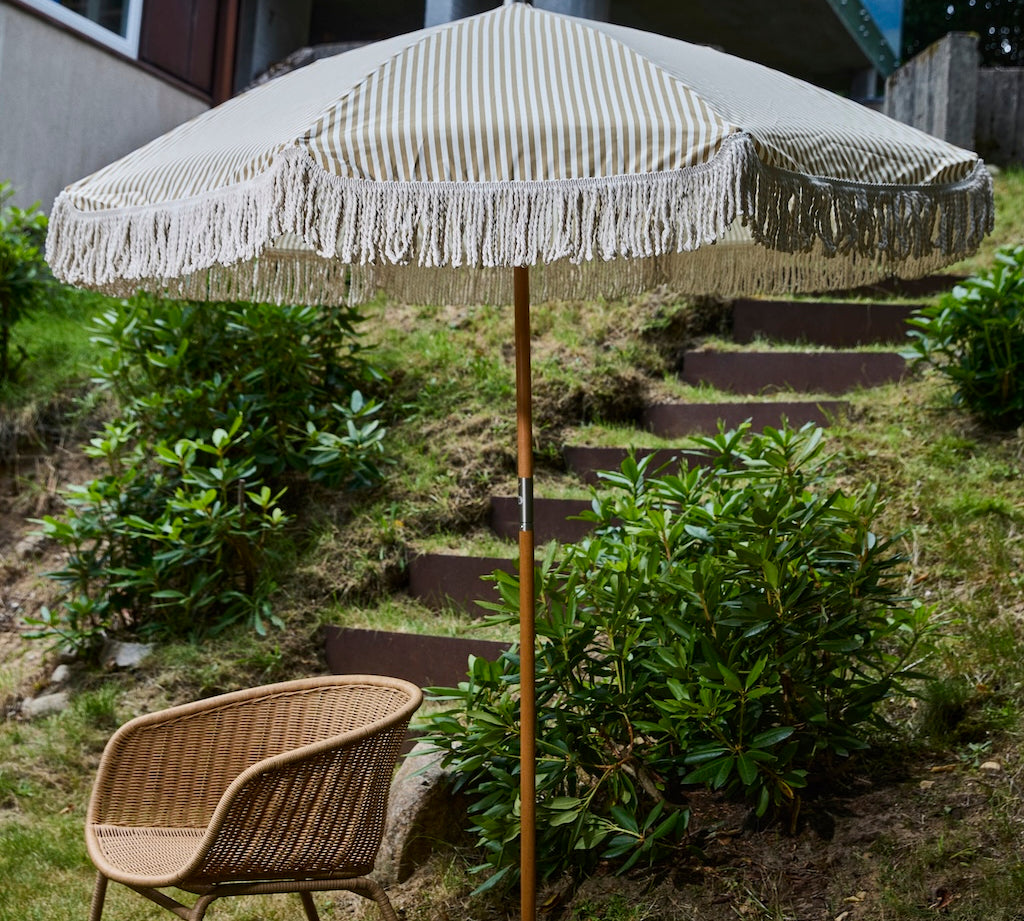 Beige striped patio umbrella with tassels on a wooden stand in a garden setting.