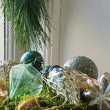 A decorative pedestal bowl made of brushed gold-toned metal, filled with green moss and assorted holiday ornaments including metallic silver and green glass baubles, displayed on a white windowsill with a pine garland in the background.
