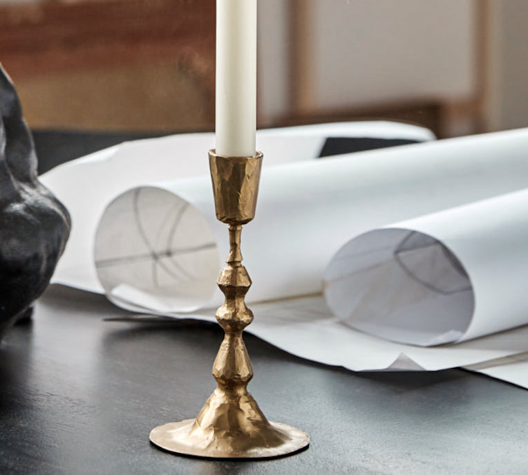 Gold candlestick with a white candle on a table with art supplies and papers.