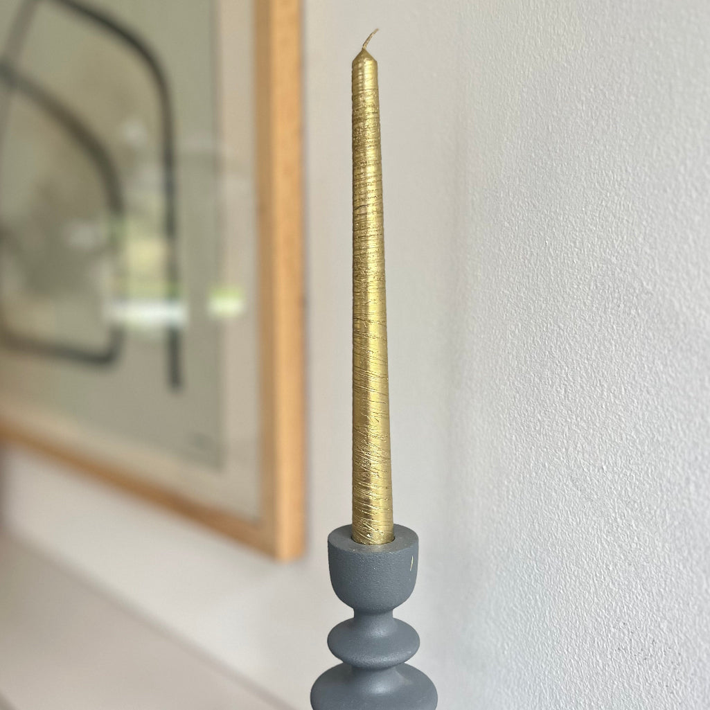 Gold candle in a gray holder against a white wall with a blurred mirror in the background