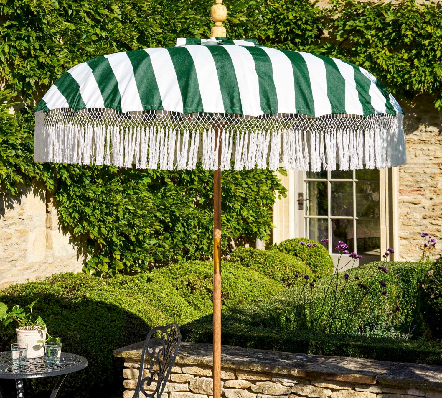 A green and white striped patio umbrella with white fringe trim stands on a stone patio beside a black metal garden table and chair. The background features a stone wall, green hedges, and a stone building with ivy.