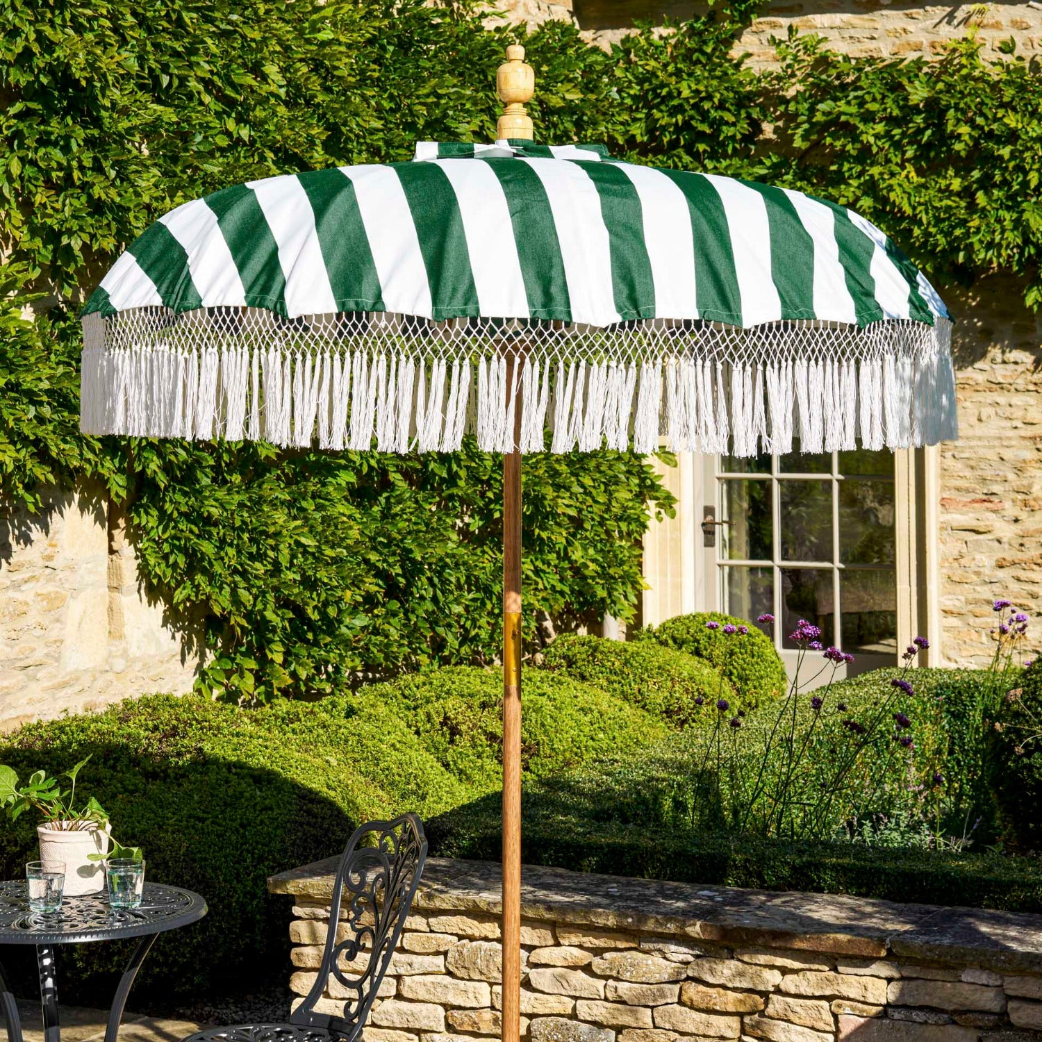 A green and white striped patio umbrella with white fringe trim stands on a stone patio beside a black metal garden table and chair. The background features a stone wall, green hedges, and a stone building with ivy.
