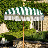 A green and white striped patio umbrella with white fringe trim stands on a stone patio beside a black metal garden table and chair. The background features a stone wall, green hedges, and a stone building with ivy.