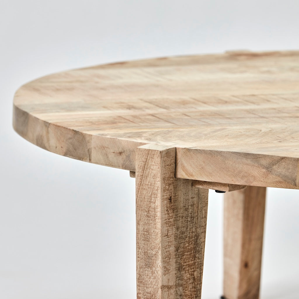Close-up of a round wooden table with a natural light beige and brown wood grain finish, showing the thick legs and joint details against a light gray background.