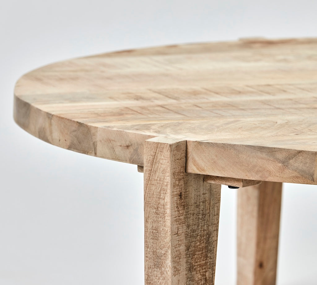 Close-up of a round wooden table with a natural light beige and brown wood grain finish, showing the thick legs and joint details against a light gray background.