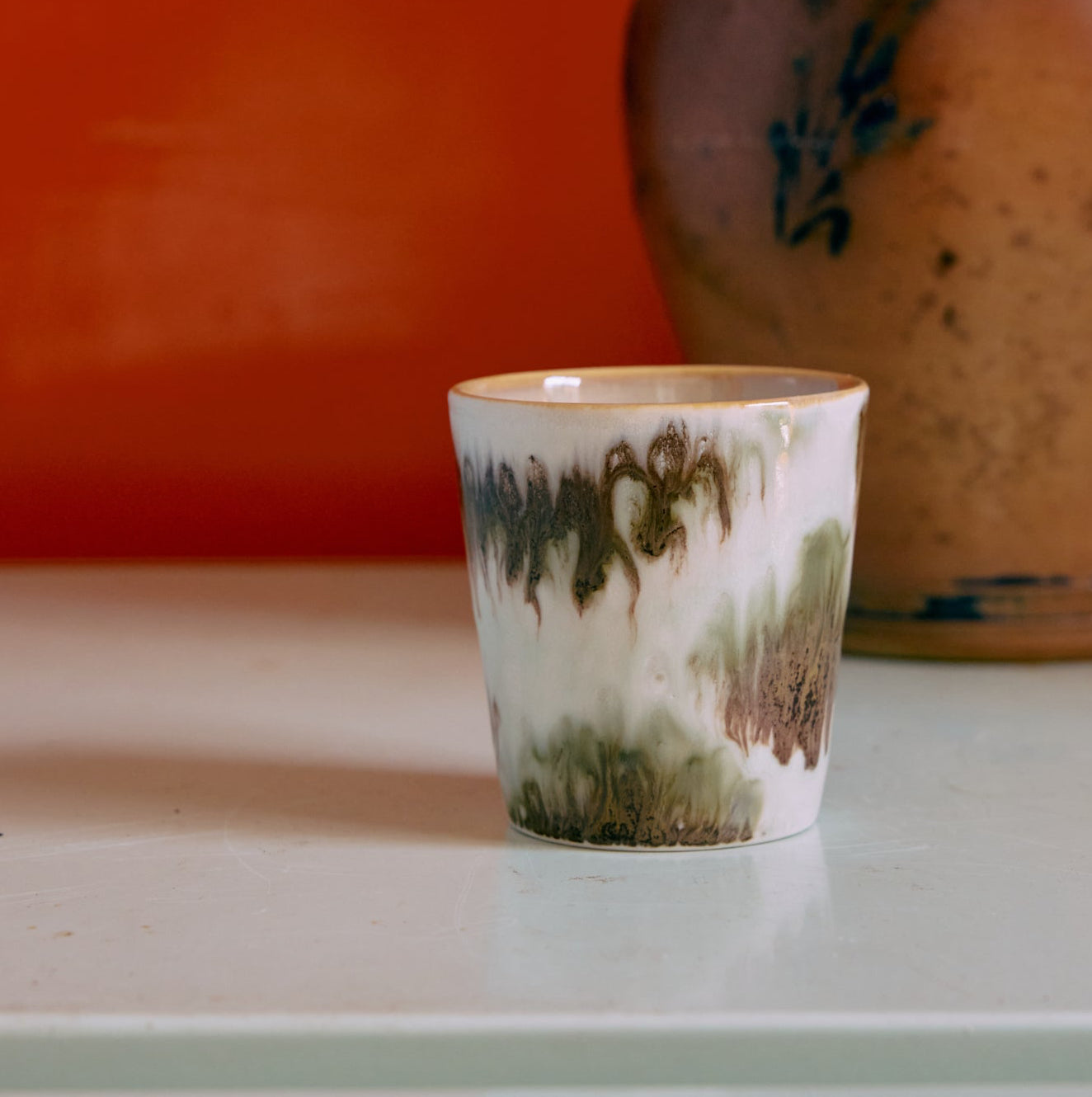 Ceramic cup with abstract green and brown design on a white surface with an orange background