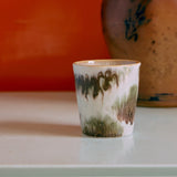 Ceramic cup with abstract green and brown design on a white surface with an orange background