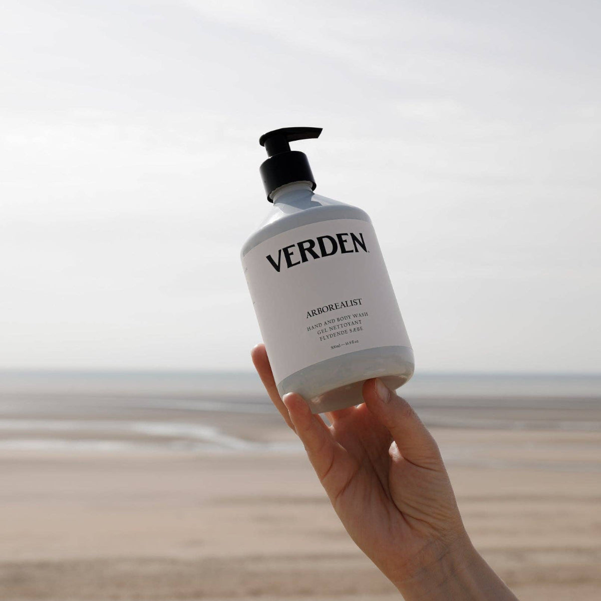 Hand holding a bottle of Verden soap with a beach in the background