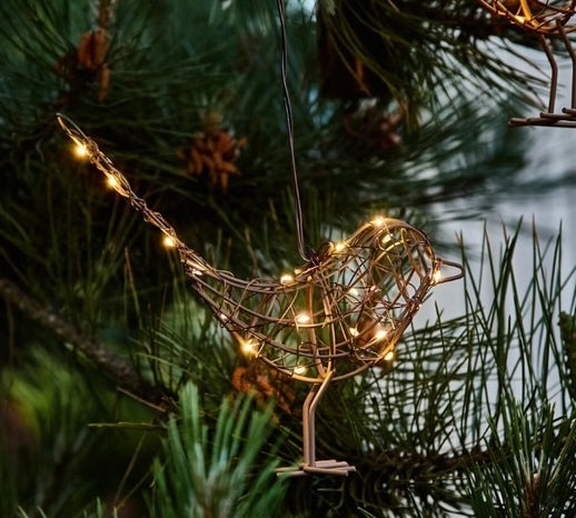 A copper-colored wire Robin Christmas ornament decorated with warm white string lights, hanging on a pine tree branch.